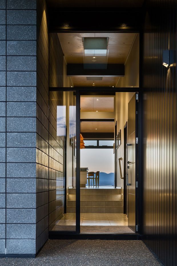 A dark, refined entrance viewed from outside, with a glazed black-framed door opening into a warmly lit, multi-level interior. The entry passage has charcoal horizontally tiled masonry on the left, vertically ribbed dark cladding on the right, a timber-lined ceiling with recessed lighting and a skylight, and tiled steps leading toward a dining area and expansive glazing with a distant mountain view.