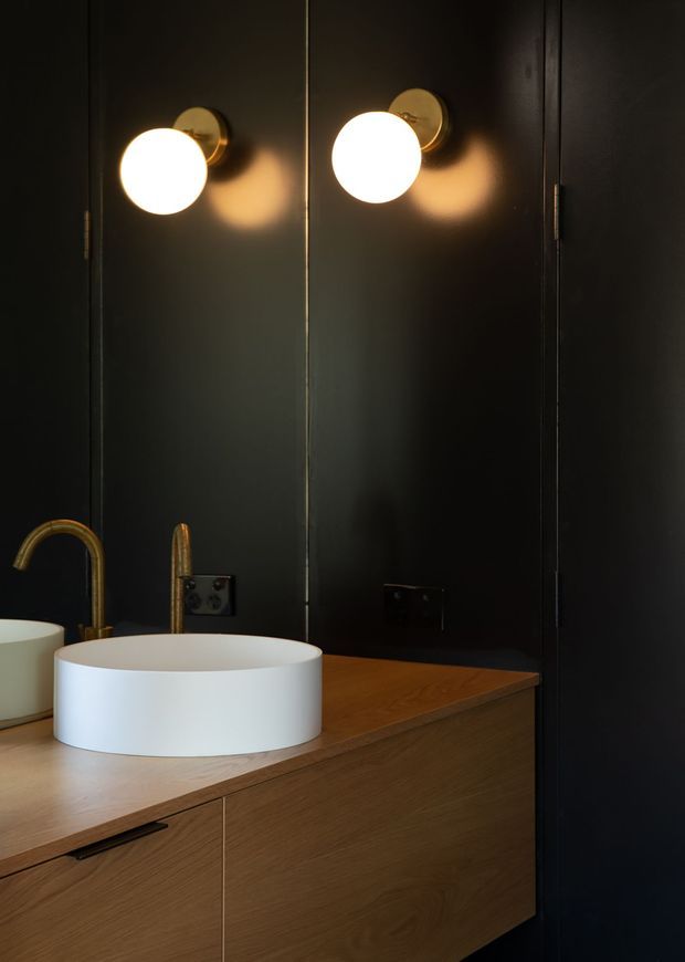 A compact, moody bathroom vanity with a round white vessel basin set on a warm timber floating cabinet. The dark wall panels and mirrored surface are accented by two glowing globe wall sconces, while brushed brass taps and hardware add warmth and contrast.