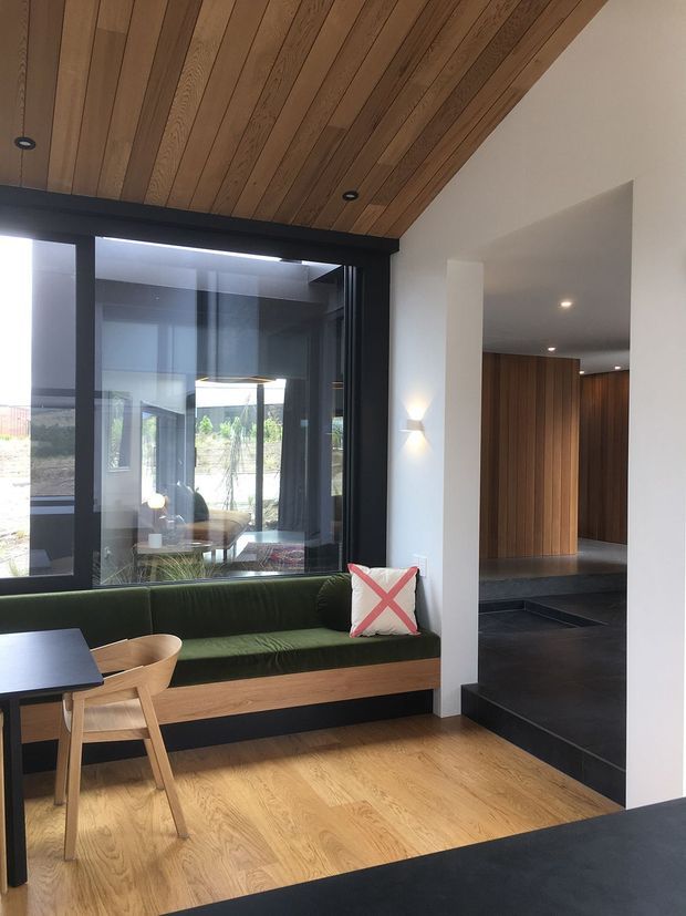 Contemporary interior featuring a built-in green upholstered window seat with timber base, a small timber dining table and chairs, black-framed glazing, and a warm timber-lined sloped ceiling. A dark tiled or stone-finished entry and hallway lead toward a vertical timber-panelled wall, while white walls and integrated lighting create a refined, modern atmosphere.
