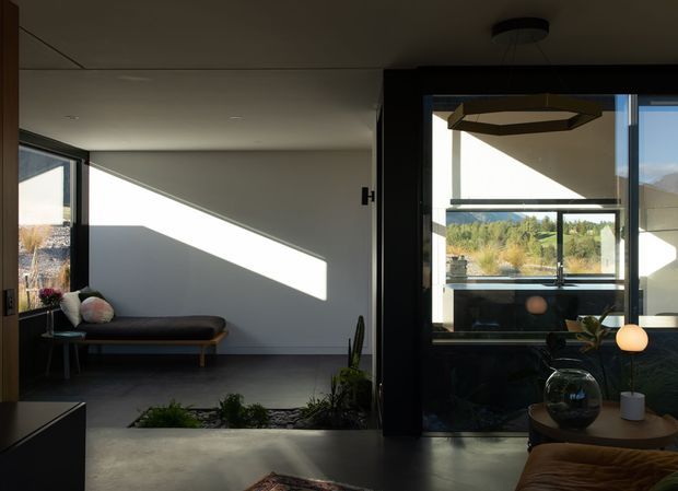 A contemporary open-plan interior with a minimalist lounge area, dark-framed floor-to-ceiling glazing, and a sunken planted courtyard or garden bed separating the seating zones. Strong angular sunlight falls across the pale walls, while an adjacent sitting area contains a low sofa, round side table, plants, and sculptural pendant lighting. The glazing looks onto a green, hilly landscape.