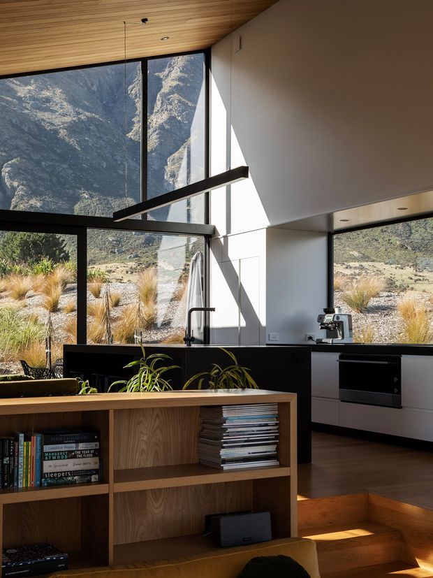 A modern open-plan kitchen and living area with dramatic floor-to-ceiling glazing overlooking rugged mountains and dry alpine vegetation. The space features a black kitchen island with a gooseneck tap, white integrated cabinetry, dark-framed windows, warm timber ceiling boards, timber flooring, built-in timber shelving, and a long linear black pendant light. Sunlight creates strong geometric shadows across the white walls, while timber steps connect the kitchen to the lower living area.