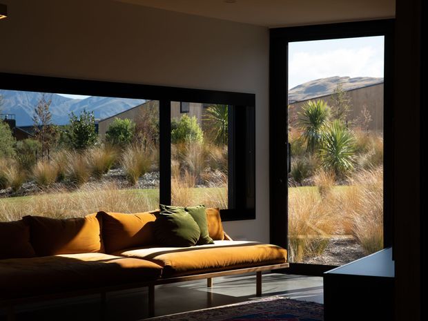 A dark, warmly lit modern living room with a low caramel-brown sofa positioned beside expansive black-framed windows and a glazed door. The room opens visually onto a landscaped dry garden with ornamental grasses and sculptural plants, with rugged hills visible beyond. The interior features a neutral ceiling and walls, dark window frames, timber-toned flooring, and a partially visible patterned rug.