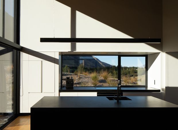 A minimalist contemporary kitchen with a large dark island and integrated sink in the foreground, extensive black-framed glazing, white built-in cabinetry, and a slim linear pendant light. Strong afternoon sunlight creates sharp geometric shadows across the walls and ceiling, while a rugged mountain landscape is visible through the window.