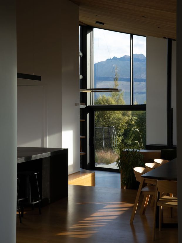 A refined open-plan kitchen and dining area with dark minimalist cabinetry, a black stone island, timber flooring, and a warm timber-lined ceiling. Full-height black-framed glazing brings in strong afternoon light and frames a dramatic mountain landscape. A dining table with timber chairs occupies the right side, while open shelving and integrated cabinetry define the kitchen zone.