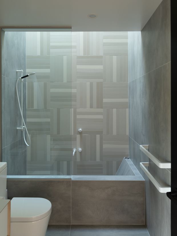 Compact contemporary bathroom with a shower-over-bath arrangement. The space features a geometric patterned pale tile feature wall, grey tiled side walls and bath surround, white sanitaryware, a handheld shower, and minimalist white towel rails.