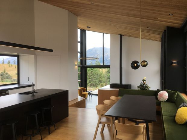 Open-plan contemporary interior combining a kitchen, dining and living area. The space features a dark kitchen island with stools, light timber flooring, white walls, extensive black-framed glazing, a timber-lined ceiling, a large dining table with timber chairs, and a green built-in sofa. Mountain and landscape views are visible through the windows.