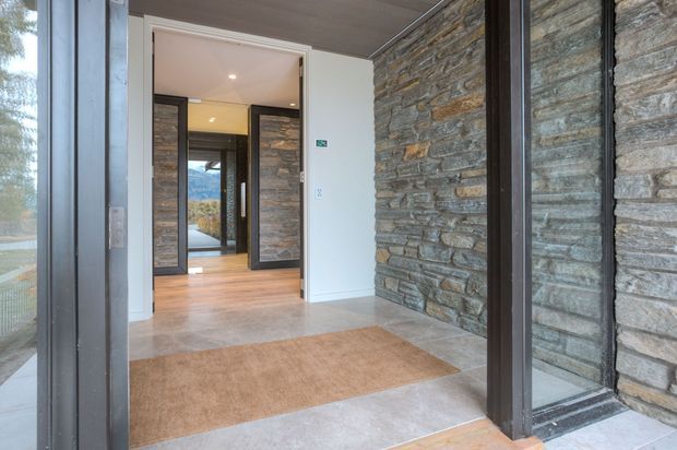 Contemporary residential entryway with a glazed dark-framed entrance, exposed stacked natural-stone wall, pale stone floor tiles, and a warm timber floor visible through the open interior doorway. The view leads into a bright hallway with additional stone detailing and exterior landscape glimpsed beyond.
