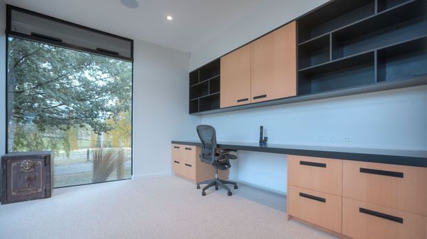 A bright, contemporary home office with a long built-in desk and extensive wall-mounted storage. Warm timber cabinetry is paired with dark open shelving and a dark worktop, while a large glazed window provides views of trees and daylight. The room also includes a wheeled office chair, pale carpet, and a compact decorative cabinet.