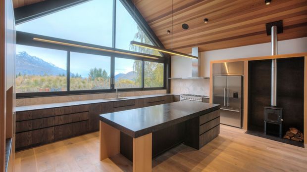 A spacious contemporary kitchen with a large central island, dark wood cabinetry, stainless-steel appliances, and a dramatic full-height corner window overlooking a mountainous landscape. The room features a warm timber-lined ceiling with recessed lighting, polished dark stone island and benchtops, a marble-look splashback, and a built-in wood-burning stove beside the refrigerator.