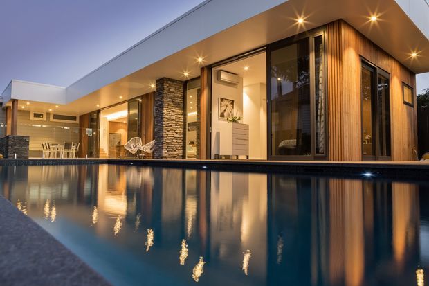 Twilight exterior view of a modern single-storey residence beside a reflective swimming pool. The home features a broad flat roof, extensive floor-to-ceiling glazing, dark metal-framed openings, warm vertical timber cladding, and a stacked-stone feature wall. Interior living and dining areas are visible through the glazing, with concealed warm lighting illuminating the covered outdoor edge.