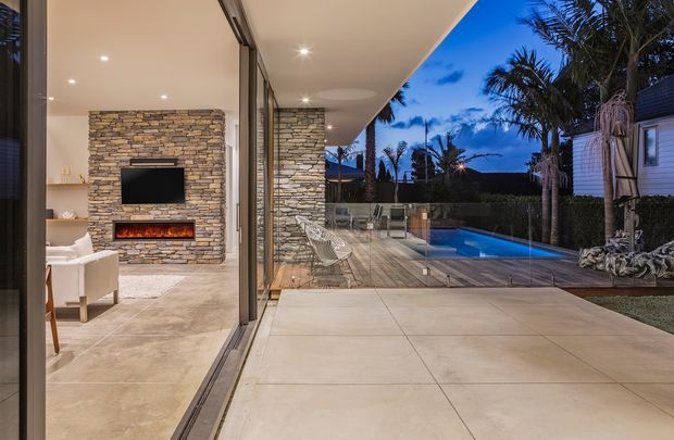 Twilight view from a contemporary indoor living area through fully open floor-to-ceiling glazing onto a tiled terrace and swimming pool. The interior features a stacked-stone fireplace wall with built-in television and linear fire, while the outdoor area includes a covered patio, glass pool fencing, palm planting, and a neighbouring coastal-style home. The scheme uses warm neutral stone and expansive indoor-outdoor connection.