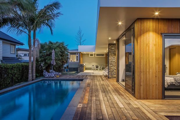 Twilight exterior view of a contemporary residential courtyard with a swimming pool, expansive timber decking, vertical timber-clad walls, stone feature columns, extensive glazing, and a covered outdoor entertaining area. A bedroom is visible through the open glazed doorway, with landscaped palms and neighboring residences in the background.
