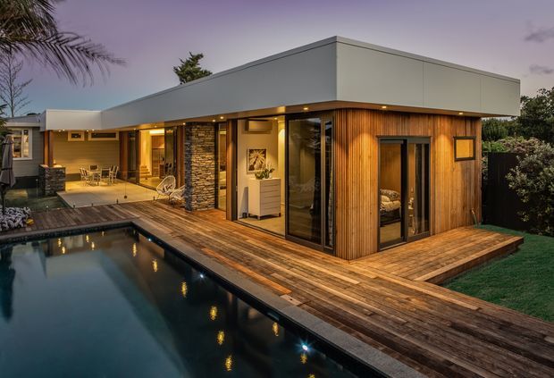 Contemporary single-storey home photographed at dusk, with a flat-roofed white upper volume, warm vertical timber cladding, dark stacked-stone feature walls, extensive glazing, and a timber deck surrounding a swimming pool. The image also shows an outdoor dining area and landscaped garden.