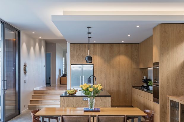 Open-plan contemporary kitchen and dining area with extensive warm timber cabinetry, a central island with sink, integrated stainless-steel refrigerator, built-in appliances, pendant lighting, and a timber dining table. Steps lead toward a hallway and glazed exterior doors are visible at the left.