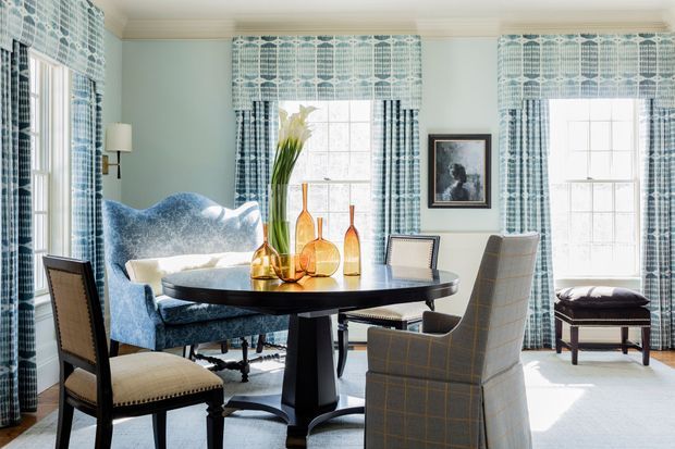 Bright, elegant dining area with a round dark dining table, upholstered dining chairs, and a large blue patterned armchair. Pale blue walls, tall windows, and matching blue-and-white patterned curtains create a refined, traditional interior. Decorative amber glass vessels and framed artwork add warm accents.