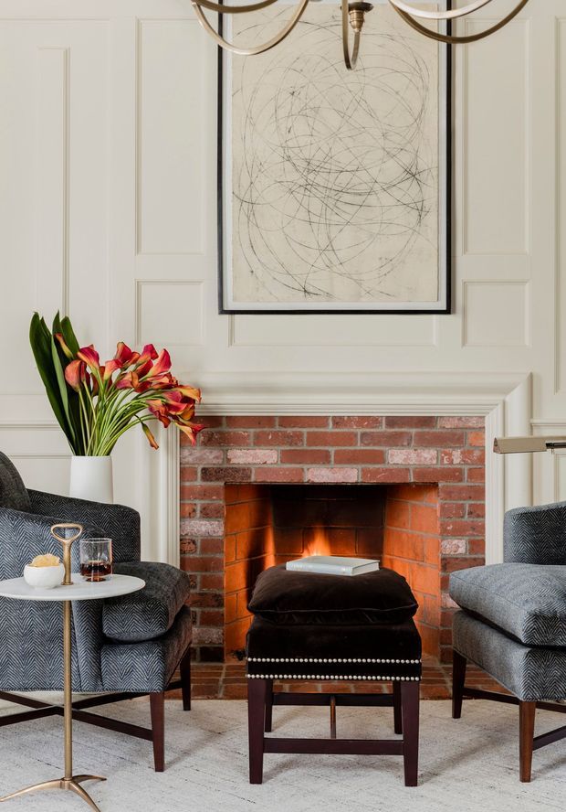 A refined sitting area centered on a traditional red-brick fireplace with a cream-painted paneled mantel wall. Two dark patterned armchairs flank a black upholstered ottoman, with a small round side table, vase of flowers, drinkware, framed abstract artwork, and a brass chandelier. The room combines classic architectural detailing with tailored, contemporary furnishings.