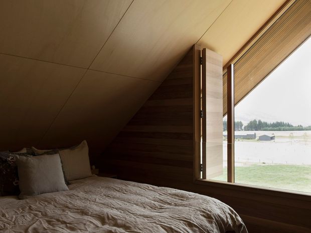 A compact, warm-toned bedroom beneath a sloping ceiling, featuring a low bed with rumpled neutral linen, dark horizontal timber wall panelling, a timber-lined ceiling, and large timber-framed glazing with a woven blind overlooking a rural landscape.