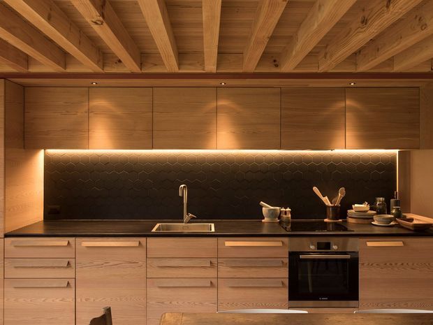 Warm, contemporary timber kitchen with extensive light-wood cabinetry, a dark stone benchtop and black hexagonal-tile splashback. Exposed timber ceiling beams and integrated warm under-cabinet lighting create a refined, intimate atmosphere. The kitchen includes a sink, cooktop and built-in oven, with open shelving and utensils arranged along the counter.