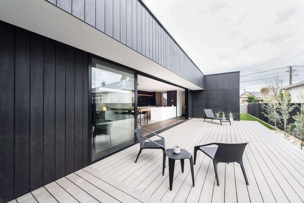 Contemporary single-storey home with dark vertical cladding, large black-framed glazing, and a spacious light timber-toned outdoor deck. The deck is furnished with modern outdoor chairs and tables and connects directly to an open-plan kitchen visible through the glazing. A small landscaped garden and neighbouring residential context appear in the background.