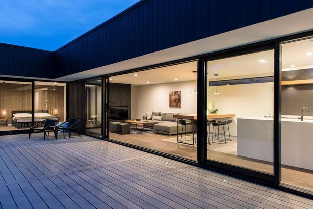 Contemporary single-level residence opening onto a spacious timber deck and courtyard. Dark vertical cladding and extensive black-framed sliding glass doors create a strong indoor-outdoor connection, with a bright open-plan living area and minimalist white kitchen visible inside. The deck includes an outdoor dining and lounge setting, while the interior features neutral furnishings, timber flooring, and recessed lighting.