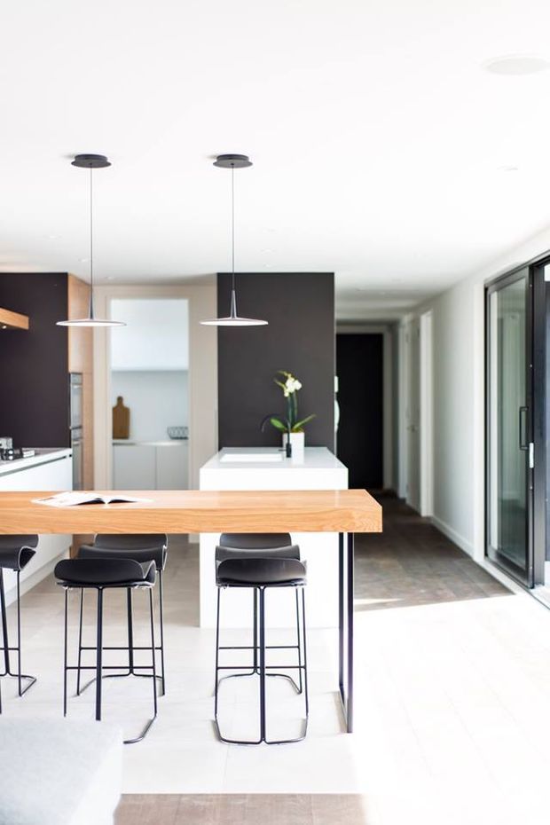 Bright, contemporary open-plan kitchen and dining area with a long light timber island bench, black bar stools, white cabinetry, dark feature cabinetry, slim pendant lights, and large glazed doors along the right side. A hallway and additional kitchen joinery are visible beyond the island.