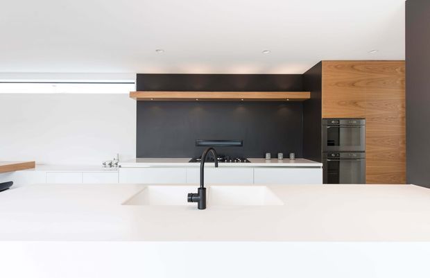 Minimalist contemporary kitchen featuring a large white island with an integrated double sink and matte black tap, white cabinetry and benchtops, a dark charcoal splashback, warm timber overhead shelving, and tall timber cabinetry housing built-in ovens. A narrow horizontal window provides natural light.