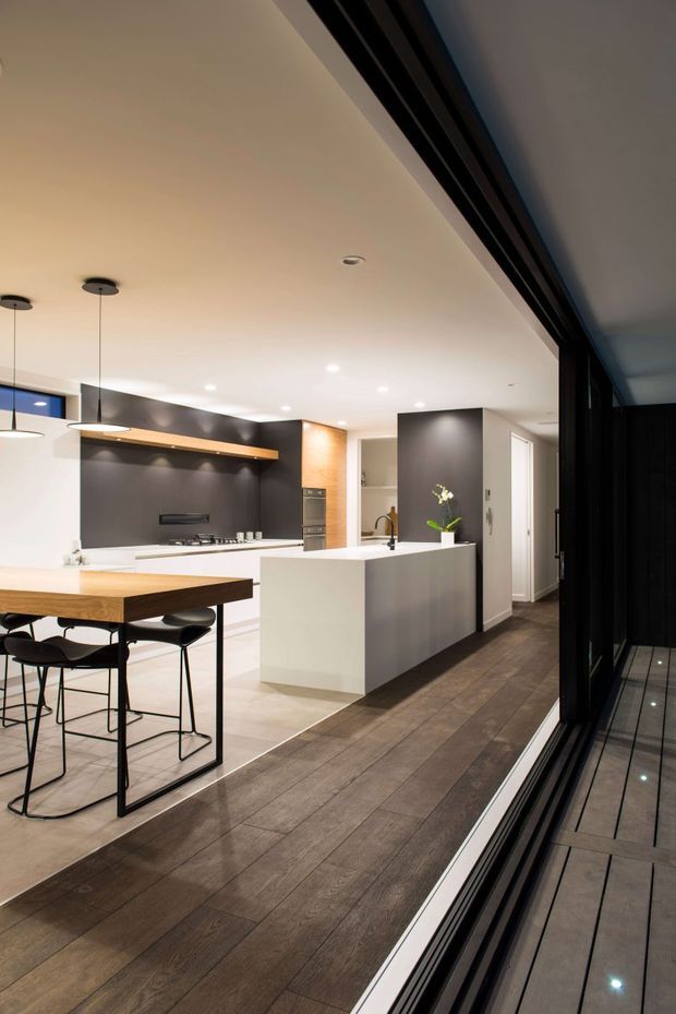 A contemporary open-plan kitchen viewed from beside large dark-framed glazing. The space features a white island with integrated sink, dark charcoal splashback and cabinetry, a timber breakfast bar with black stools, pendant lighting, recessed ceiling lights, and wide dark timber flooring. A hallway and utility area are visible beyond the kitchen.