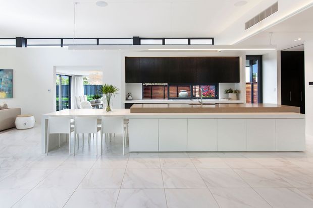 Bright, open-plan contemporary kitchen and living area featuring a large white island with integrated cabinetry, a pale stone benchtop, white tiled flooring, and a dark timber feature wall surrounding the television and kitchen display shelving. Floor-to-ceiling glazing and open doors connect to an outdoor area, while linear black-framed glazing and recessed ceiling lighting add modern architectural detail.