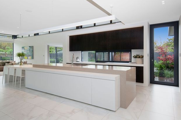 Bright, open-plan contemporary kitchen with a large white island, pale stone benchtops, integrated cabinetry, and a dramatic dark timber feature wall. The space includes a dining area, extensive glazing, skylight-like ceiling detailing, and a glazed door opening to a planted outdoor area.