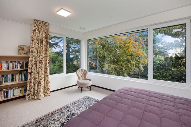 Bright bedroom with large corner windows overlooking leafy trees, a mauve quilted bed in the foreground, a vintage upholstered chair, built-in or freestanding wooden bookshelves, patterned curtains, and a traditional rug. The room has white walls and ceiling with simple contemporary detailing.
