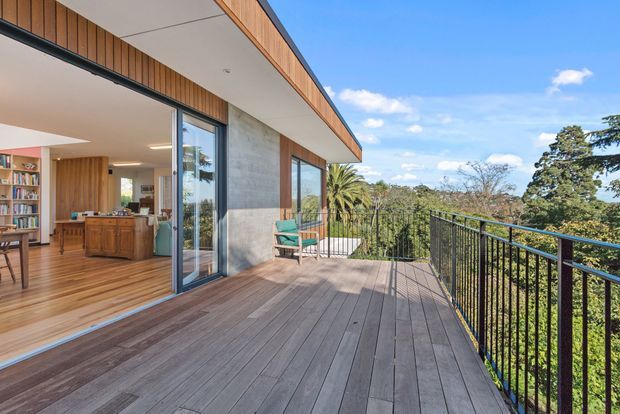 A contemporary home exterior with a large timber deck and black metal balustrade overlooking a leafy landscape. Full-height sliding glass doors open to a warm timber-lined interior with a visible kitchen, dining area, and bookshelf. The building features a low-pitched projecting roof, timber cladding, exposed concrete walls, and expansive glazing.