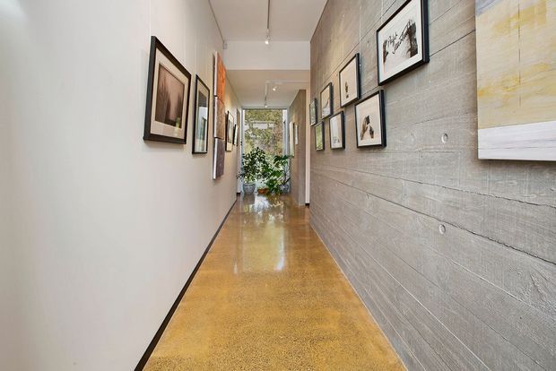 A long, narrow contemporary hallway with polished warm-toned concrete flooring, a smooth white wall on the left, and a textured grey concrete feature wall on the right. Framed artwork is arranged along both walls, with a glass door and indoor plants visible at the far end. Recessed track-style ceiling lighting illuminates the gallery-like corridor.