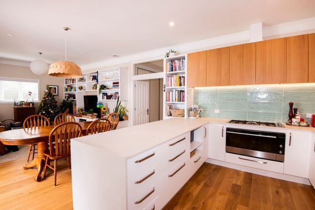 Bright open-plan kitchen and dining area with a large white island, white lower cabinetry, warm timber upper cabinets, a pale green textured tile splashback, and integrated stainless-steel cooking appliances. The adjacent dining area has a timber table and chairs, pendant lighting, timber flooring, and built-in shelving with books and decor.