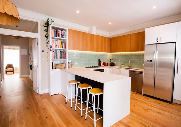 Bright contemporary kitchen with warm timber cabinetry, white island and tall cabinetry, stainless-steel appliances, pale green textured splashback, and timber flooring. The island includes seating for three, with an open hallway and built-in shelving visible to the side.