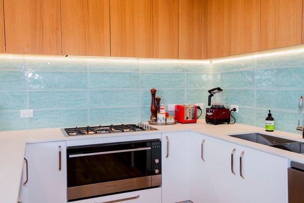 A compact L-shaped kitchen with white lower cabinetry and warm timber overhead cabinets. The kitchen features a pale aqua tiled splashback, integrated oven, gas cooktop, corner benchtop, stainless-steel sink, under-cabinet lighting, and small countertop appliances.