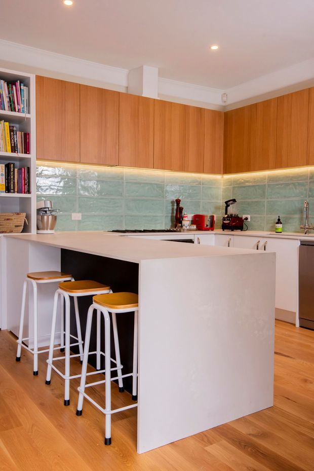 A bright, contemporary kitchen with a large white island incorporating seating for three, flat-front timber cabinetry, pale aqua-green glazed tile splashback, white benchtops, open shelving, and integrated under-cabinet lighting. Warm timber flooring and colourful small appliances add contrast.