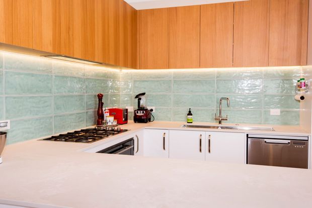 A compact L-shaped kitchen with warm timber overhead cabinetry, white lower cabinets, a pale stone-look benchtop, and glossy pale aqua tiled splashback. The kitchen includes a gas cooktop, oven, sink, dishwasher, small appliances, and integrated under-cabinet lighting.