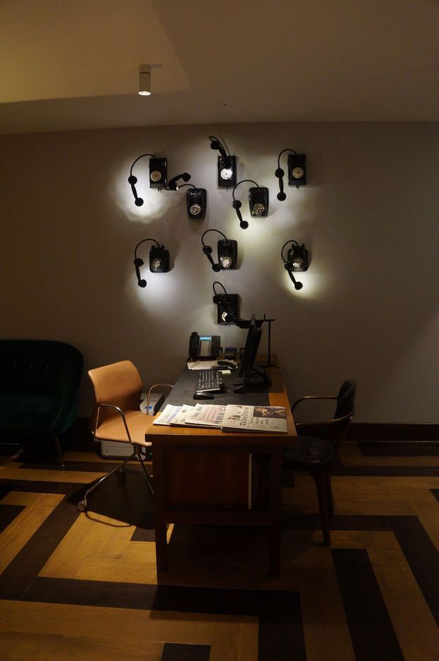 Dimly lit, vintage-inspired workspace with a dark wood desk, computer equipment, office chairs, and a striking arrangement of old black wall-mounted telephones used as decorative lighting. The room has warm beige walls, recessed ceiling lighting, and a geometric parquet-style timber floor with dark inlaid bands.