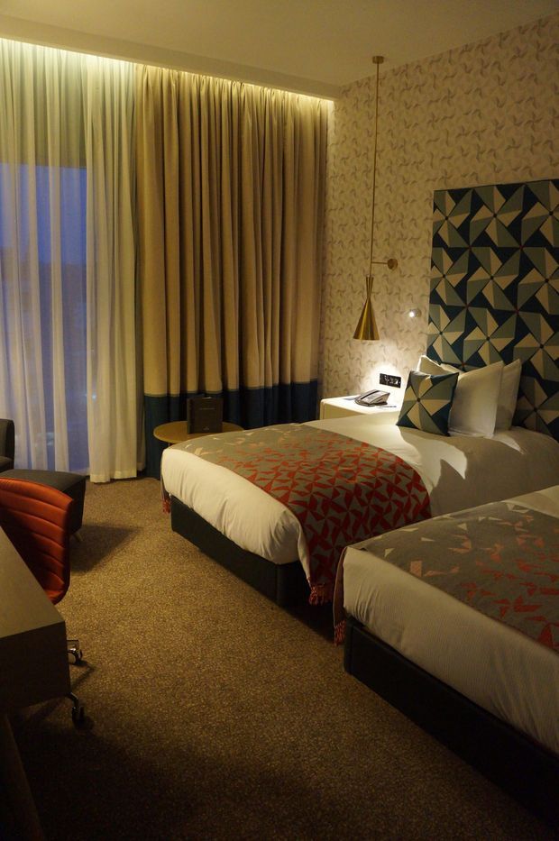 A warmly lit contemporary hotel-style bedroom with two beds, floor-to-ceiling layered curtains, patterned carpet, geometric upholstered headboards, and coordinated geometric bed throws. A small bedside table with a conical pendant light sits beside the main bed, while a desk and chair are partially visible in the foreground.
