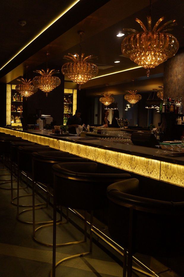 A dark, luxurious bar interior with a long illuminated counter, black upholstered bar stools, mirrored walls, bottle displays, and multiple ornate amber-gold glass chandeliers shaped like floral globes. Warm accent lighting and reflective surfaces create a dramatic upscale atmosphere.