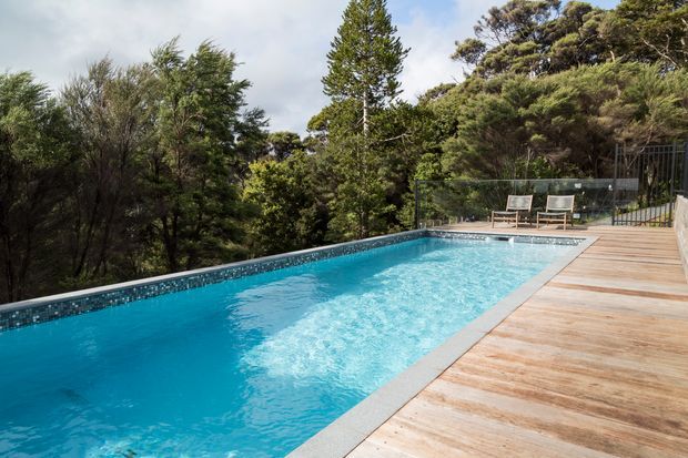 A long outdoor swimming pool surrounded by dense mature vegetation, with a timber deck along one side and a glass safety balustrade at the far end. Two lounge chairs sit beside the pool, creating a private, landscaped recreational setting.