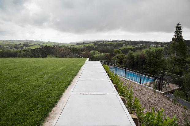A broad concrete pathway runs along the edge of a manicured grassy lawn, overlooking a fenced swimming pool and landscaped grounds. Rolling green hills and scattered homes are visible in the distance beneath an overcast sky.