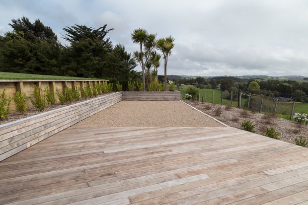 An expansive timber deck and gravel terrace overlooking a rural green landscape. The terrace is bordered by tiered horizontal timber retaining walls with climbing plants, fencing, and sculptural palm-like trees under an overcast sky.
