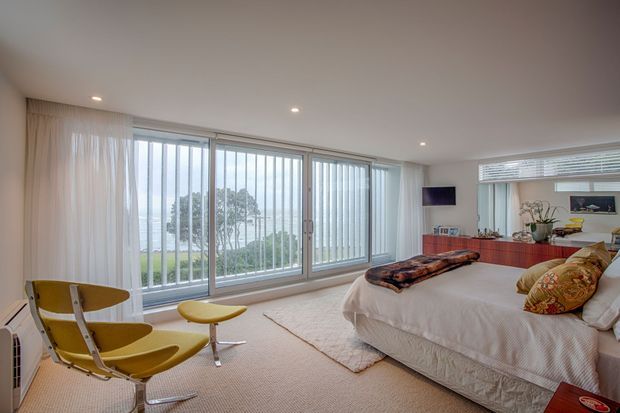 Bright contemporary bedroom with a large curved wall of floor-to-ceiling glazing, vertical blinds and sheer curtains overlooking greenery. The room contains a low-profile upholstered bed with patterned accent cushions, a textured neutral rug, two mustard-yellow lounge chairs, recessed ceiling lights, and a long red-toned built-in cabinet beneath a wall-mounted television and mirror.