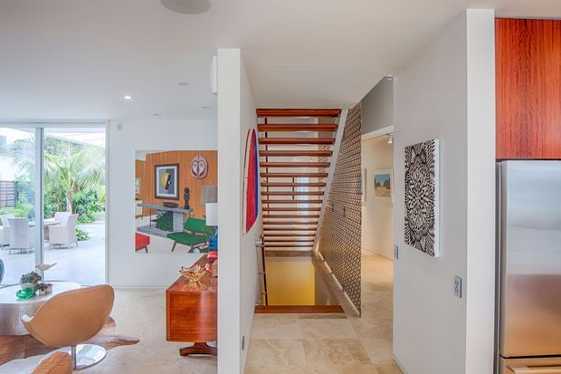 Bright contemporary open-plan interior featuring a floating timber staircase with open risers and a slim metal mesh balustrade. The staircase connects to a lower level, with white walls, pale stone flooring, timber cabinetry, artwork, and glimpses of a furnished living and dining area with tropical greenery beyond the glazing.