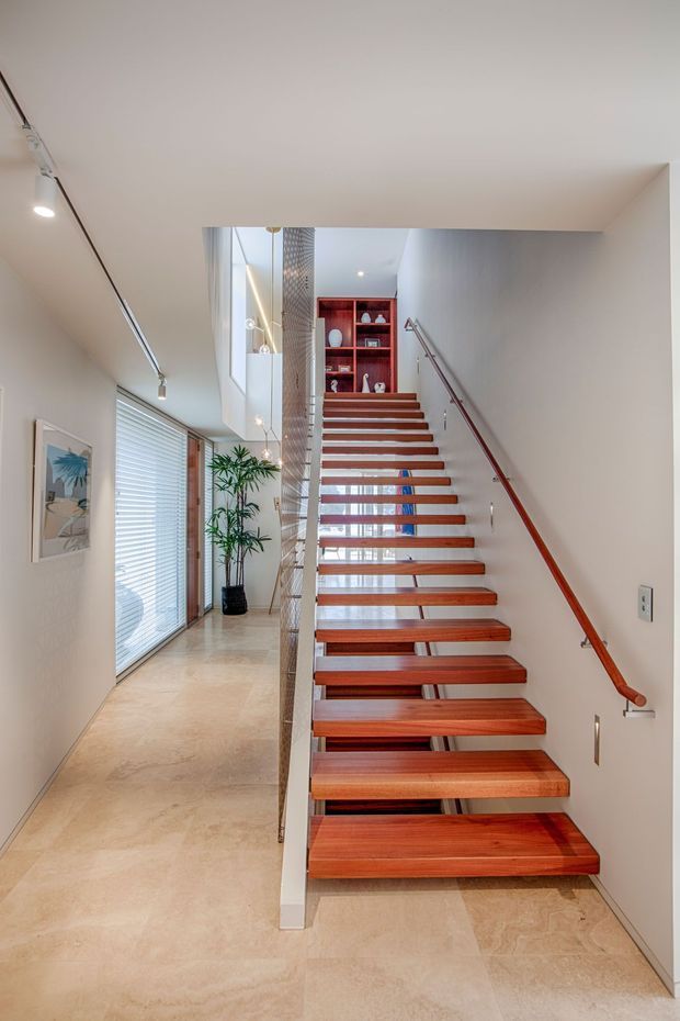 Bright contemporary interior focused on an open-riser staircase with warm timber treads, a slim metal handrail, and a glass balustrade. The stairway rises beside a light-filled hallway with large glazed openings and horizontal blinds, pale stone flooring, white walls, recessed lighting, and a red built-in display niche at the upper landing.