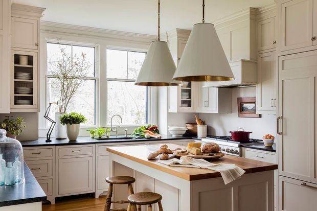 Bright, traditionally detailed kitchen with cream painted cabinetry, framed glass-front cupboards, dark countertops, a timber-topped island with stools, and large windows above the sink. Two oversized pendant lights hang over the island, while bread and cooking items provide a lived-in domestic feel.