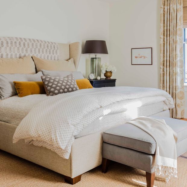 A warmly styled bedroom featuring a large upholstered bed with a patterned headboard, layered cream and taupe bedding, mustard and patterned accent pillows, and a matching upholstered footstool. A dark bedside table with a glass lamp sits beside the bed, while patterned curtains frame a bright window. The room has a soft neutral palette with natural light and refined traditional detailing.