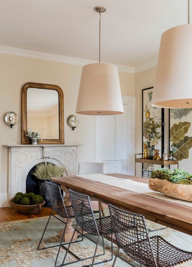 A refined dining room with a long natural wood dining table, woven black dining chairs, and oversized pendant lights. The room features a carved white fireplace surround, ornate dark wood-framed mirror, wall sconces, built-in cabinetry, decorative shelving, artwork, and warm neutral walls with traditional crown moulding. A patterned rug and indoor plant arrangements add texture and softness.