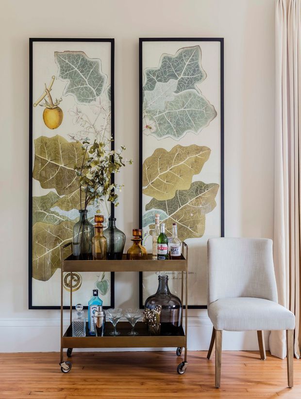 A refined interior vignette featuring a vintage-style dark wood and metal bar cart stocked with glass bottles and barware, positioned against a light wall. Two tall botanical prints in dark frames create a strong vertical backdrop, while a cream upholstered dining or accent chair sits beside floor-length curtains. Warm timber flooring and muted natural tones give the scene an elegant, inviting character.